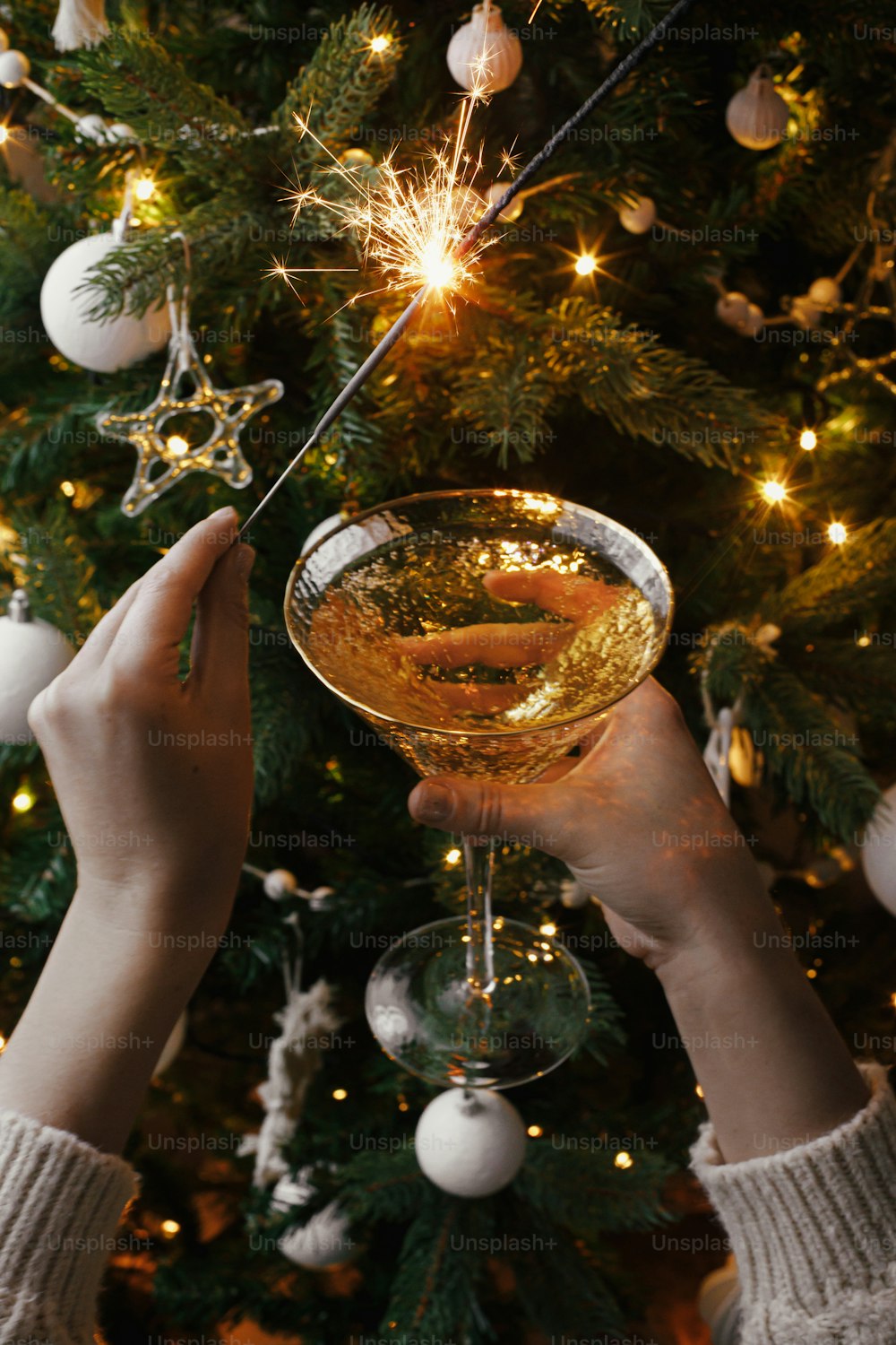 Bonne Année! Mains tenant un cierge magique brûlant et un verre de champagne sur fond de lumières d’arbre de Noël. Feu d’artifice bengale lumière et boisson dans les mains des femmes. Moment atmosphérique. Fête de la Saint-Sylvestre