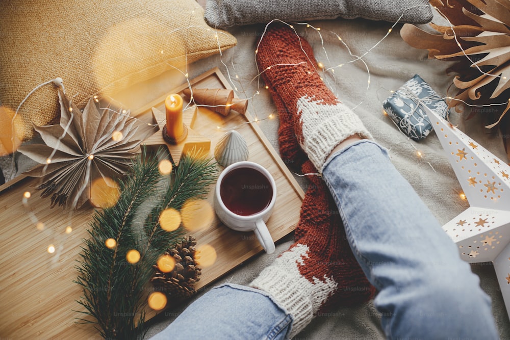 Woman feet in woolen socks on soft bed with warm cup of tea, christmas stars, golden lights, trees, candle and pillows. Top view. Cozy moments at home. Girl relaxing in scandinavian evening room