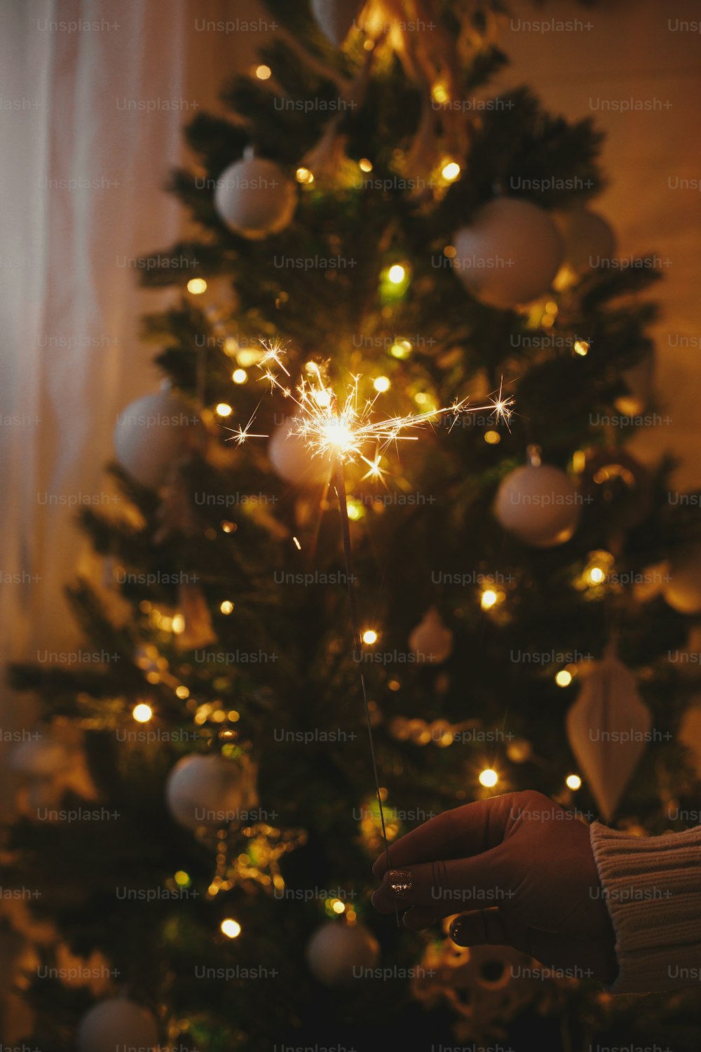 Mão segurando um brilho ardente no fundo das luzes da árvore de Natal no quarto escuro festivo. Feliz Ano Novo! Fogos de artifício bengala brilhando na mão da mulher. Espaço para texto. Momento atmosférico