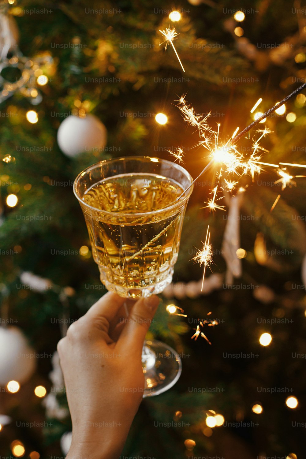 크리스마스 트리 조명을 배경으로 샴페인 잔에 불꽃놀이 빛으로 축하하는 여자. 새해 복 많이 받으세요! 스칸디나비아 방에서 불타는 폭죽과 음료를 들고 있는 손. 대기 순간