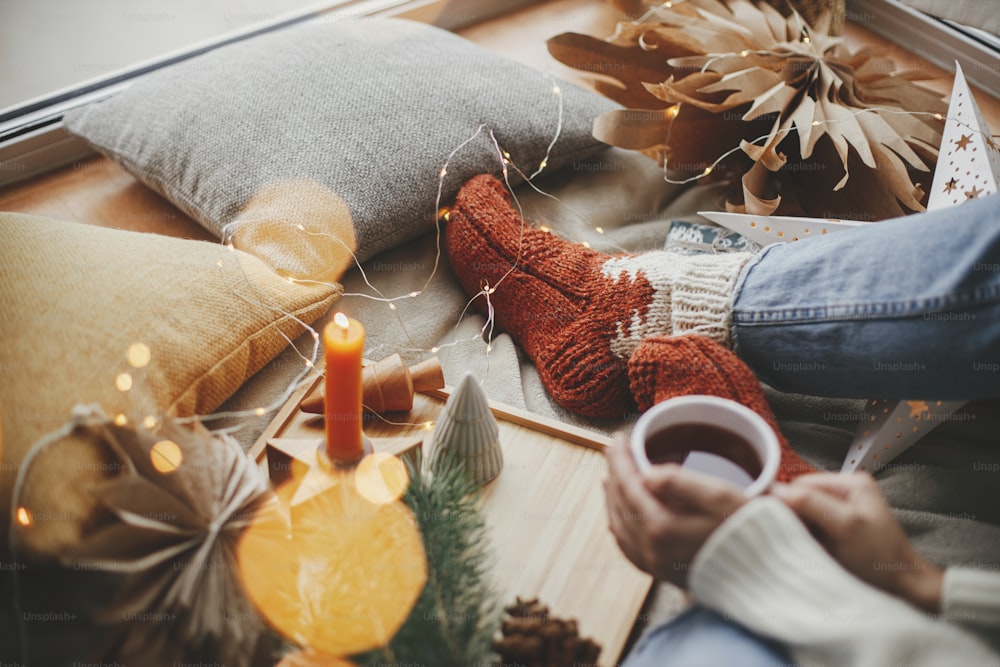Woman hands with warm cup of tea relaxing on soft bed with golden lights, christmas stars, pine, candle and pillows in scandinavian room. Atmospheric moment. Cozy winter holidays at home.
