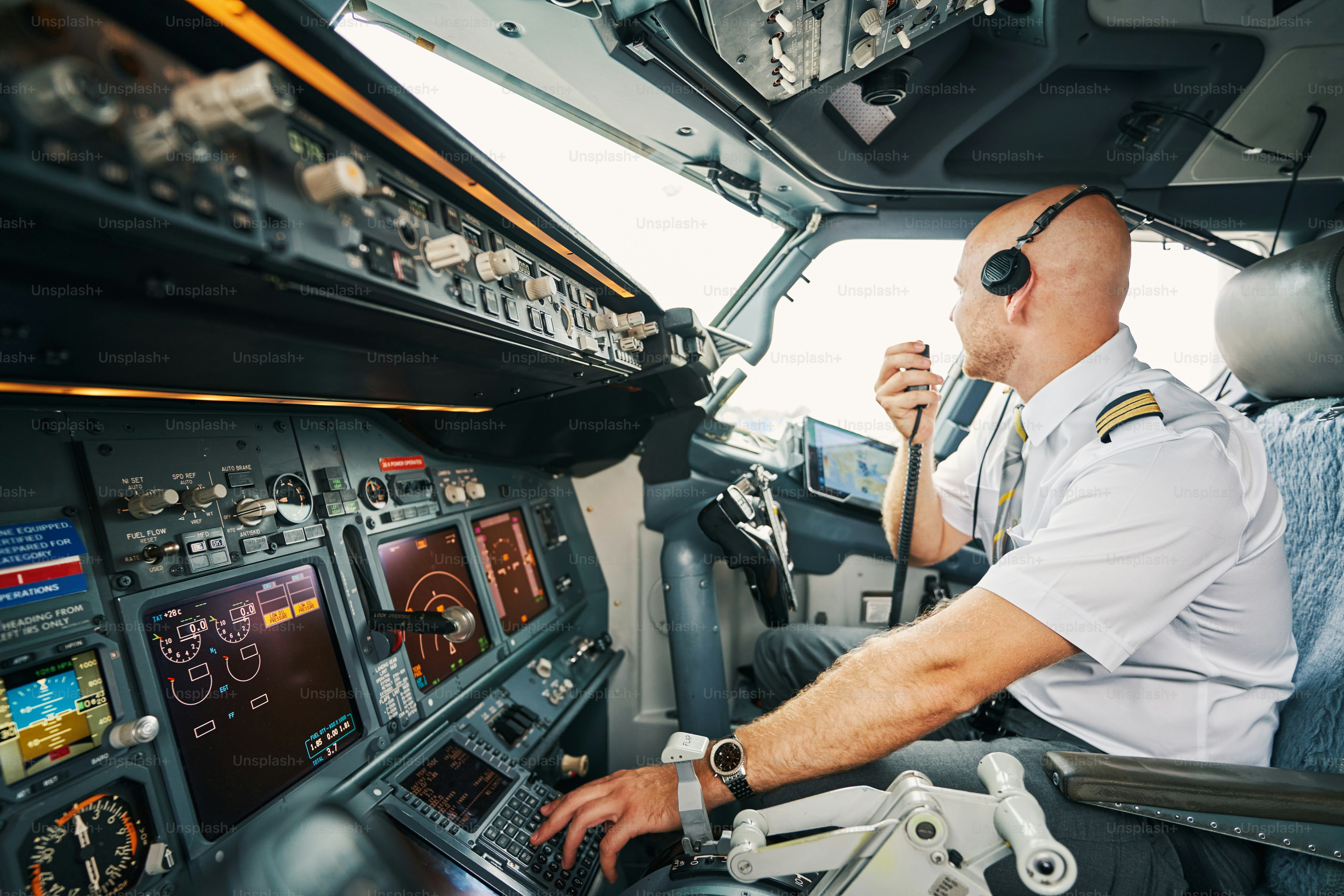 Professional male pilot in headphones seated in his cockpit with a radio in his hand looking away