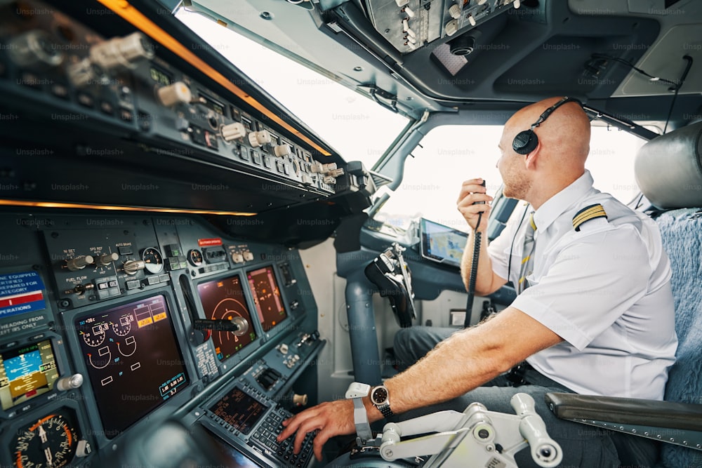Professional male pilot in headphones seated in his cockpit with a radio in his hand looking away