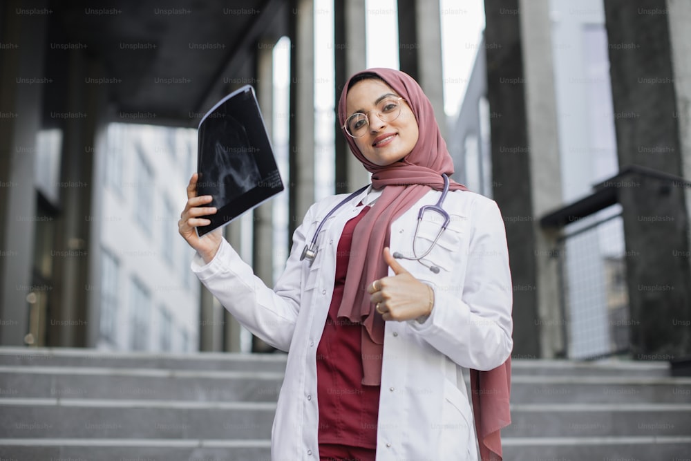 Attraktive, glückliche muslimische Ärztin im Hijab, die eine Röntgenaufnahme des Schädels des Patienten untersucht und mit Daumen nach oben in die Kamera lächelt. Arabische Ärztin im Freien mit Röntgenscan