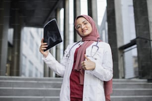 Attractive happy Muslim female doctor in hijab, examining an x-ray of patient's skull and smiling at the camera with thumb up. Arabic woman physician standing outdoors with xray scan