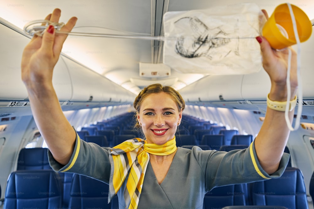 Smiling lovely young stewardess with an oxygen mask in her hands standing aboard the airplane