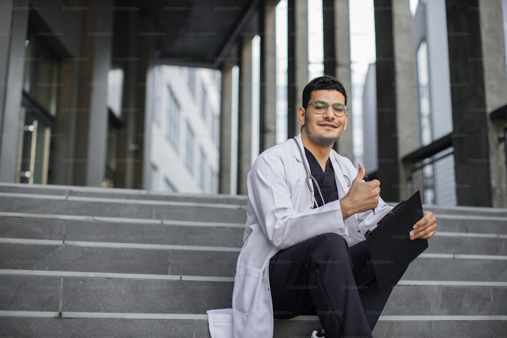 Close up horizontal shot of smiling Indian man physician, wearing scrubs and medical coat, sitting with clipboard on modern stairs outdoors and showing like to camera. Copy space