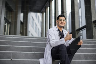 Gros plan horizontal d’un médecin indien souriant, vêtu d’une blouse et d’une blouse médicale, assis avec un presse-papiers sur des escaliers modernes à l’extérieur et montrant comme à la caméra. Espace de copie