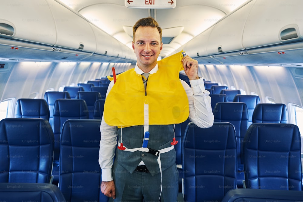 Vista frontal de um mordomo profissional sorridente em um colete salva-vidas de pé a bordo da aeronave durante a demonstração de segurança pré-voo