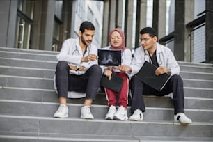 Medical team of three diverse Arabian doctors surgeons, sitting on the stairs outdoors of modern hospital and looking at skull X-ray, discussing ways of treatment and operation