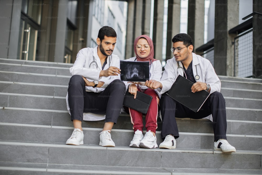 Équipe médicale de trois chirurgiens médecins arabes, assis sur les escaliers à l’extérieur de l’hôpital moderne et regardant la radiographie du crâne, discutant des moyens de traitement et d’opération