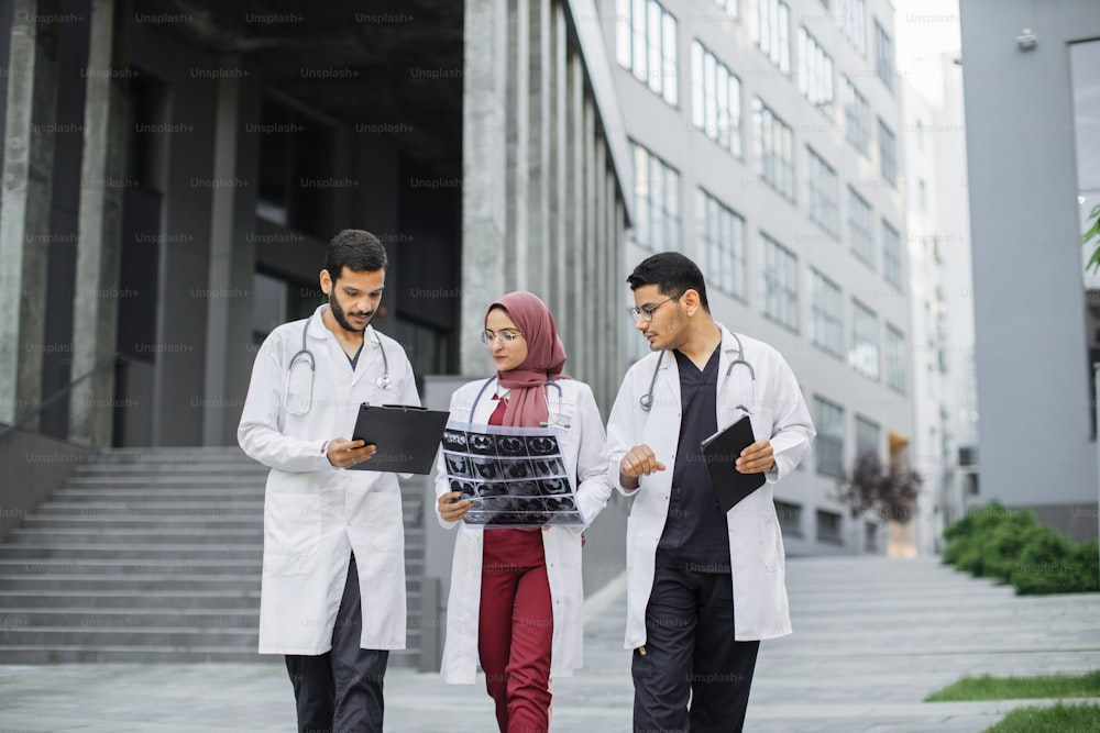 Três médicos, uma mulher de hijab e dois homens em roupas médicas, discutindo a tomografia de raios-x do paciente, andando do lado de fora no fundo do hospital moderno com escadas