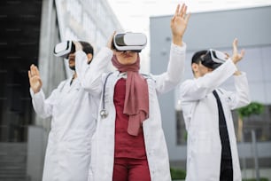 기술 및 직업 개념. VR을 사용한 시각화 교육. VR 헤드셋을 착용하고 공중에서 손을 움직이며 현대적인 병원 건물 밖에 서 있는 세 명의 집중된 아시아 의사
