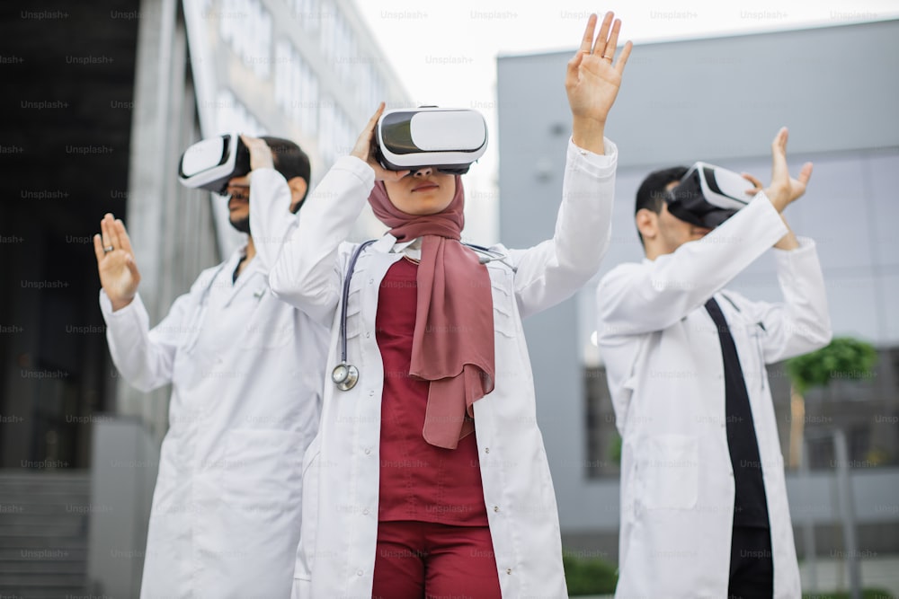 Technology and occupational concept. Visualization training with VR. Three focused Asian doctors, wearing VR headset, moving hands in the air, standing outside the modern hospital building
