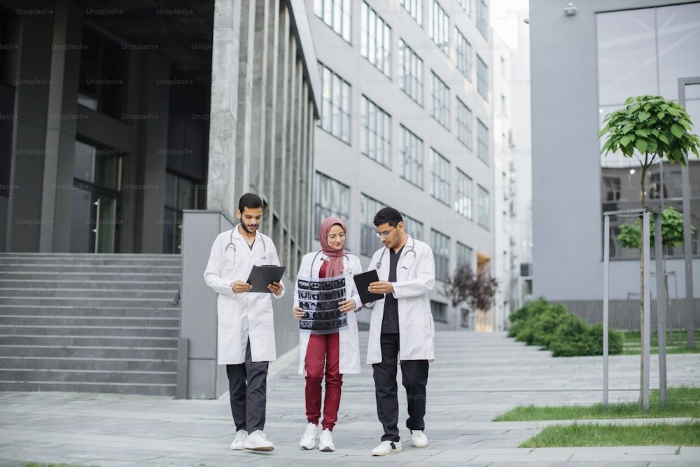 Three Arabian Muslim doctors, in medical suits and coats, discussing patient's x-ray tomography scan, walking outside on the background of modern hospital with stairs