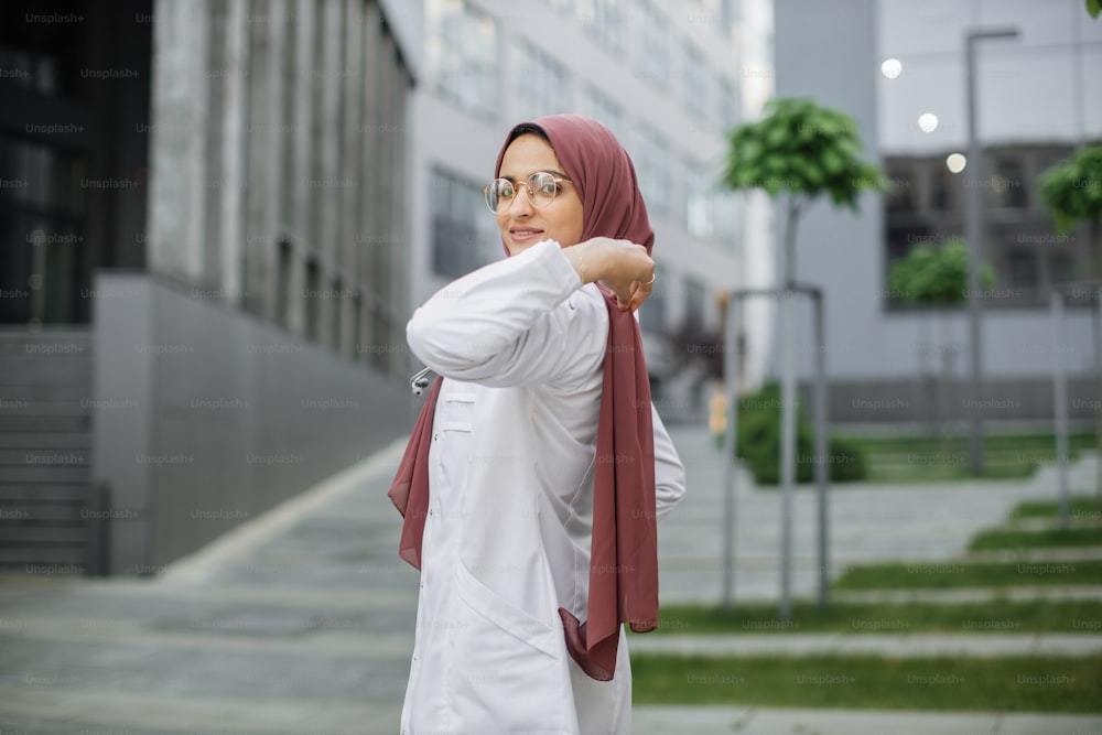 Medizin und Gesundheitswesen. Porträt einer hübschen saudi-arabischen muslimischen Ärztin oder Krankenschwester im weißen Kittel mit Stethoskop, die ihren Hijab zurechtrückt und in die Kamera schaut. Outdoor-Porträt der arabischen Ärztin