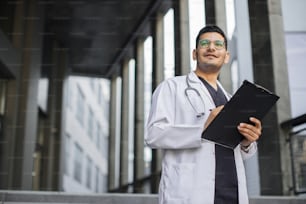 クリップボードにRX処方箋を書いている物思いにふけるプロの男性アラブ人医師の肖像画を接写します。男性医師は、近代的な診療所の屋外に立って医療フォームに記入しています。コピースペース