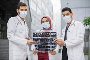 Un equipo diverso de jóvenes científicos médicos, médicos neurólogos, vistiendo uniforme y máscara, posando al aire libre cerca del hospital y mirando tomografía computarizada, resonancia magnética.