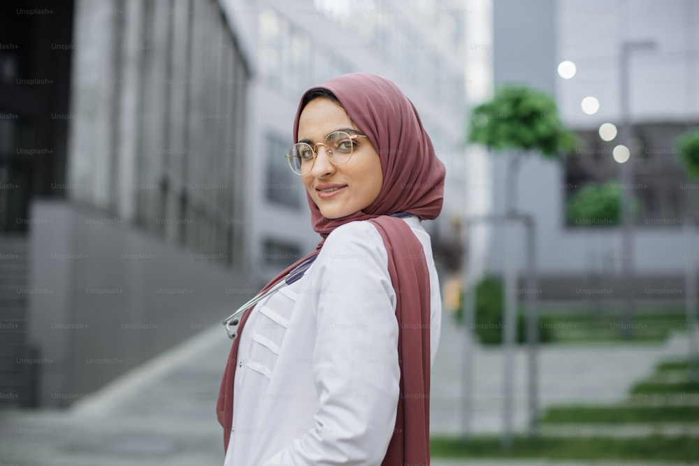 Mitarbeiter im Gesundheitswesen im Nahen Osten. Nahaufnahme des Porträts einer l�ächelnden, selbstbewussten muslimischen Krankenschwester oder Ärztin, die Hijab trägt und im Freien vor modernen Gebäuden vor der Kamera posiert
