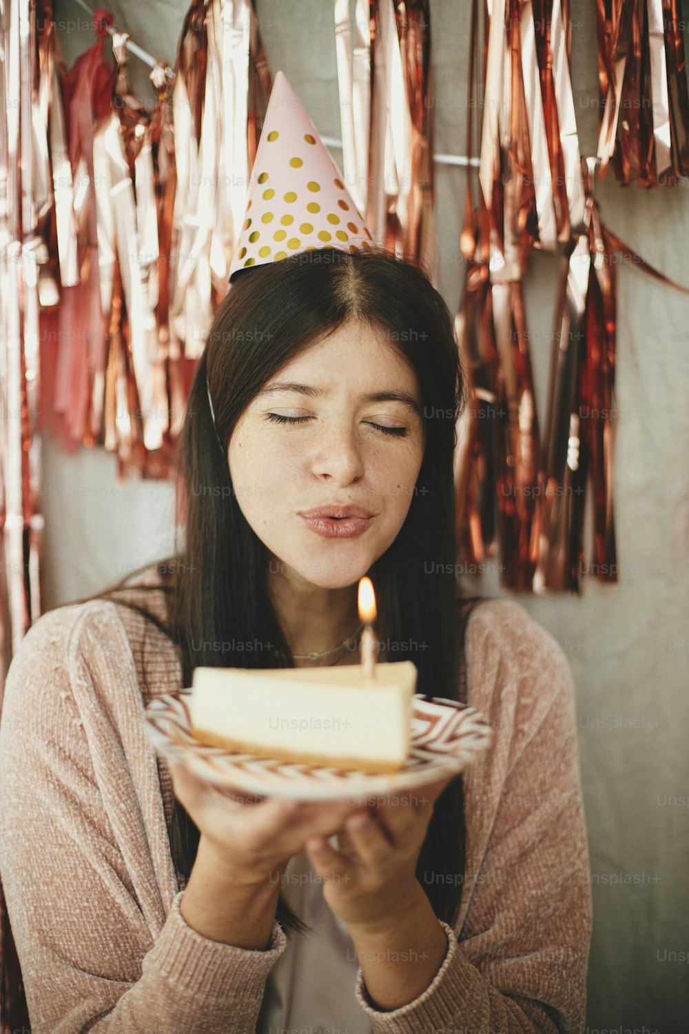 Stylish happy woman in party hat blowing burning candle on piece of birthday cake and making a wish on background of modern rose gold tassel garland in room. Celebrating birthday at home