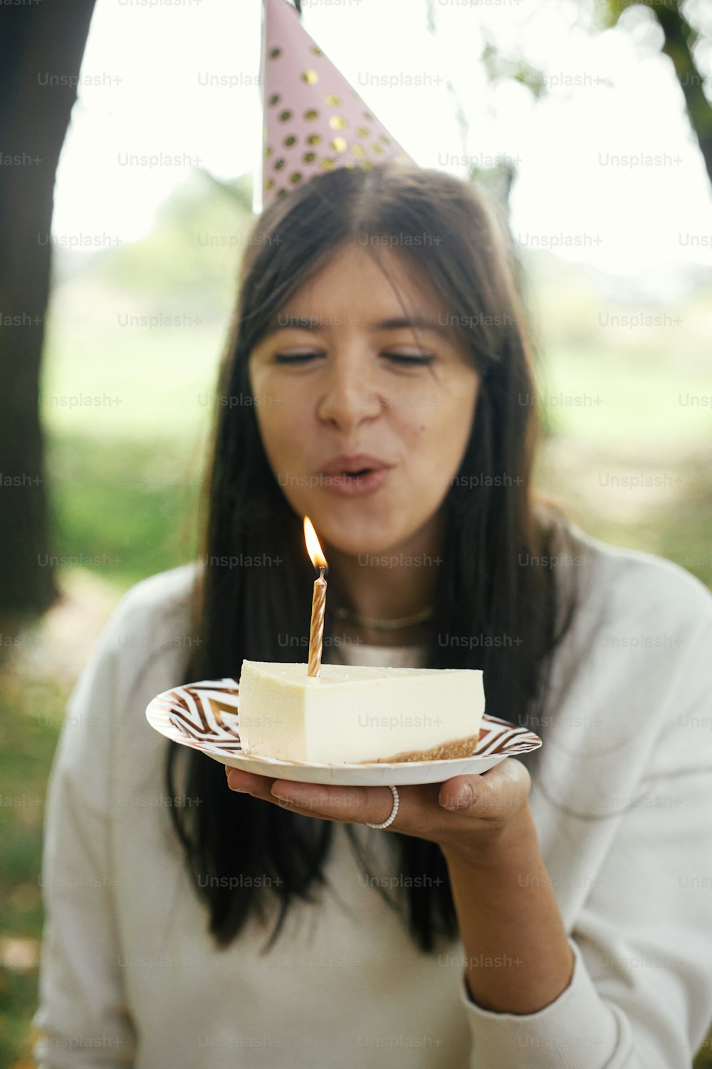 Stylish happy woman in party hat holding piece of birthday cake with burning candle and making a wish. Celebrating birthday at picnic party outdoor.