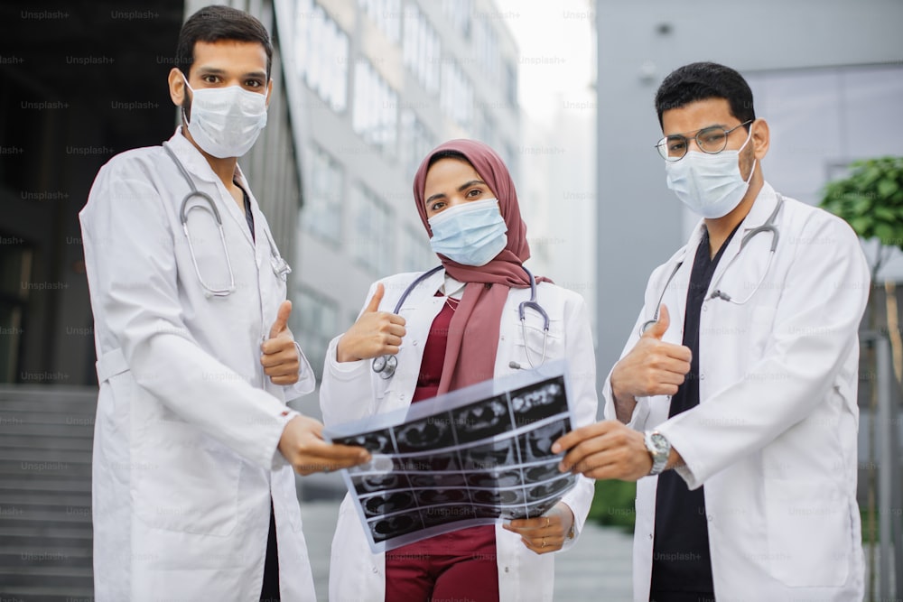 Team of three young diverse experts doctors in uniforms scrubs and protective masks, holding MRI x-ray tomography picture in front of modern clinic outdoors, looking at camera with thumbs up