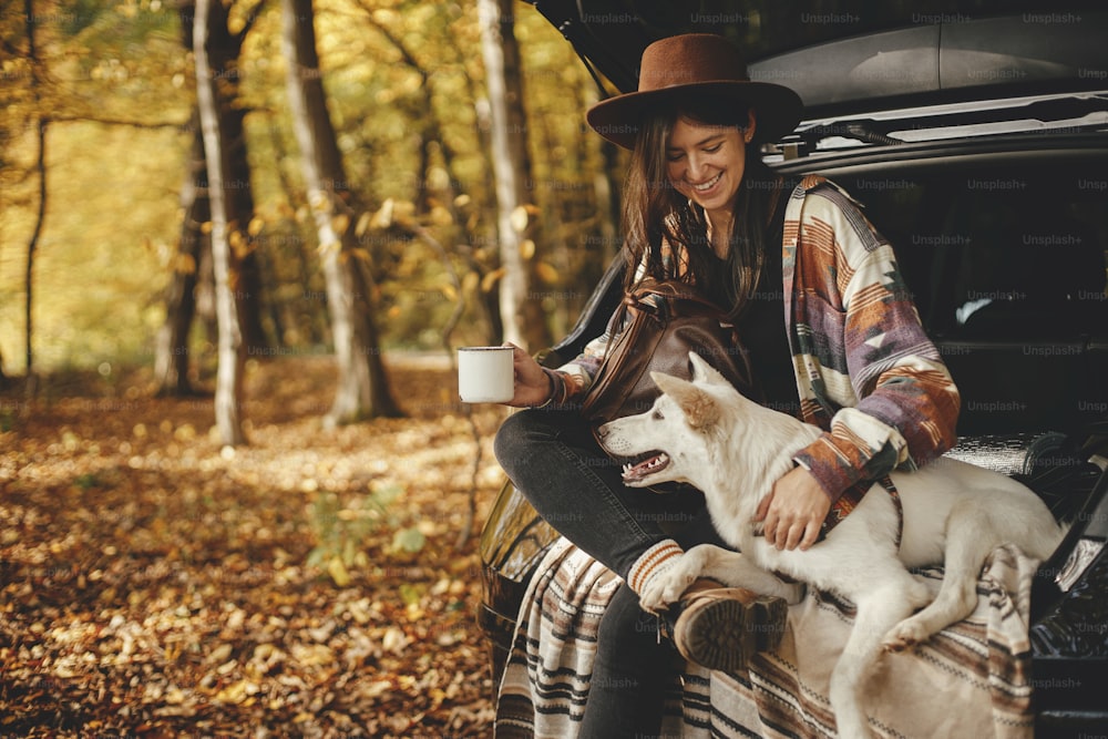 Mulher hipster elegante com copo e mochila sentada com cão bonito no porta-malas do carro em bosques ensolarados do outono. Viagem e road trip com animal de estimação. Espaço para texto. Viajante feminino novo que abraça o cão branco doce
