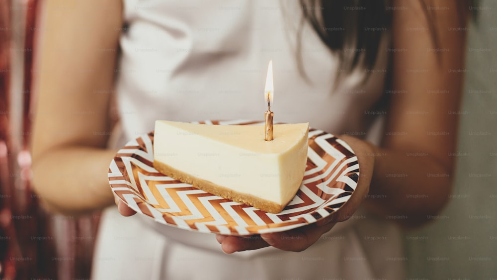 Make a wish. Piece of modern birthday cake with burning candle in hand on background of happy woman in stylish dress and modern rose gold tassel garland in room. Celebrating birthday at home