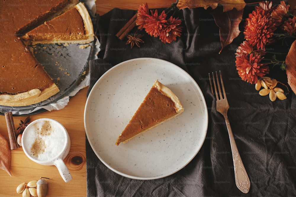 Pumpkin pie slice on modern plate and hot coffee on rustic table with linen napkin, autumn flowers and leaves, anise and cinnamon. Homemade pumpkin tart recipe. Top view. Happy Thanksgiving