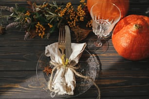 ビンテージのカトラリー、リネンナプキン、ハーブ、グラスを木製のテーブルに並べたモダンなプレート、カボチャ、日光の下での秋の花のアレンジメント。素朴な農家の結婚式。感謝祭のディナーテーブルセッティング