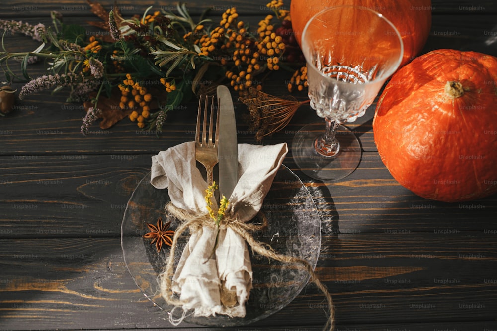 Moderner Teller mit Vintage-Besteck, Leinenserviette, Kräuter und Glas auf Holztisch mit Kürbissen und Herbstblumenarrangement im Sonnenlicht. Rustikale Bauernhaushochzeit. Thanksgiving-Tischdekoration