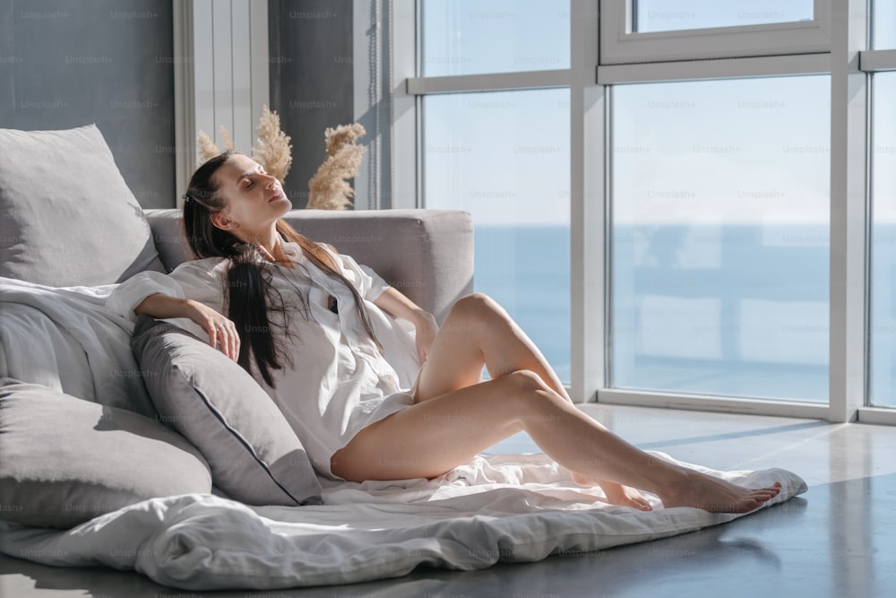 Young beautiful european woman in white shirt with naked legs leaning on sofa with white blanket and many decorative pillows, keeping eyes closed and enjoying happy morning at home after waking up