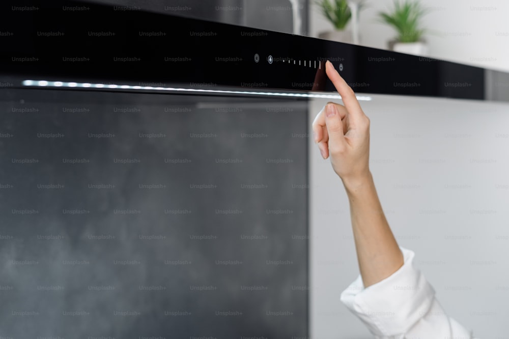 Cropped shot of female adjusting cooker hood or exhaust while cooking in minimalistic fully furnished kitchen with modern integrated appliances