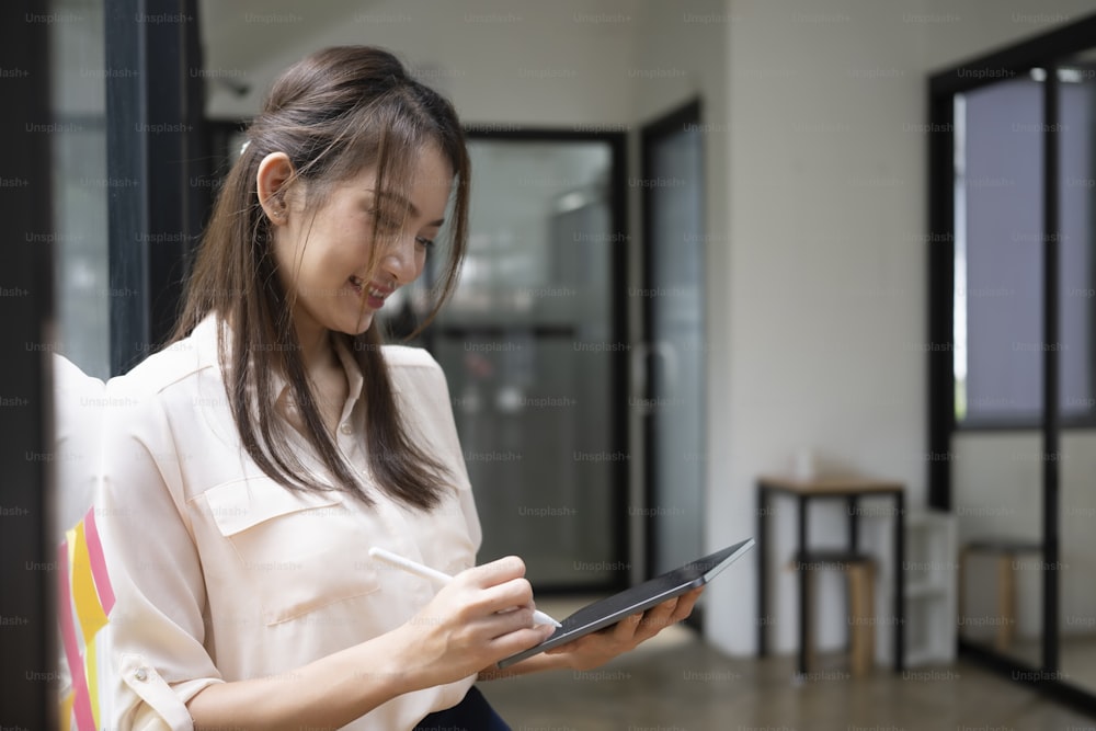 Junge Asiatin, die im Büro steht und ein digitales Tablet benutzt.