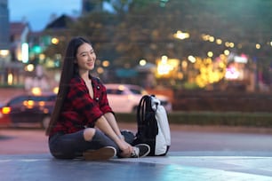 Beautiful female solo traveller sitting on stairs of shopping mall with backpack, blurred street night market in background