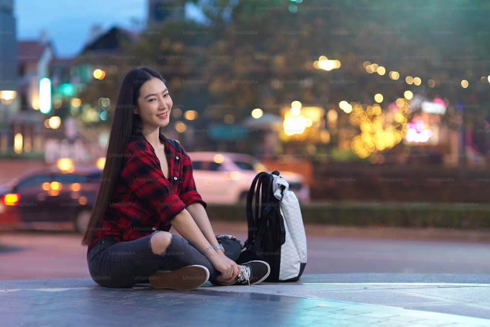 Beautiful female solo traveller sitting on stairs of shopping mall with backpack, blurred street night market in background