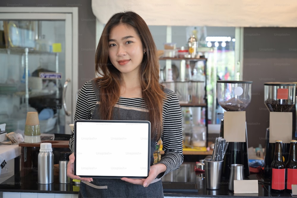 Young female entrepreneur in apron showing digital tablet while standing in modern cafe.