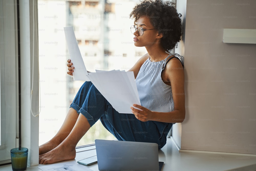 Serious female engineer in spectacles sitting on a wide windowsill with floor plans in her hands