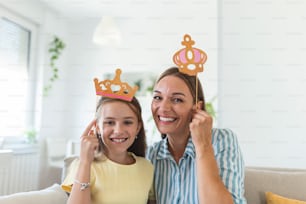 Funny family on a background of bright wall. Mother and her daughter girl with a paper accessories. Mom and child are holding paper crown on stick.