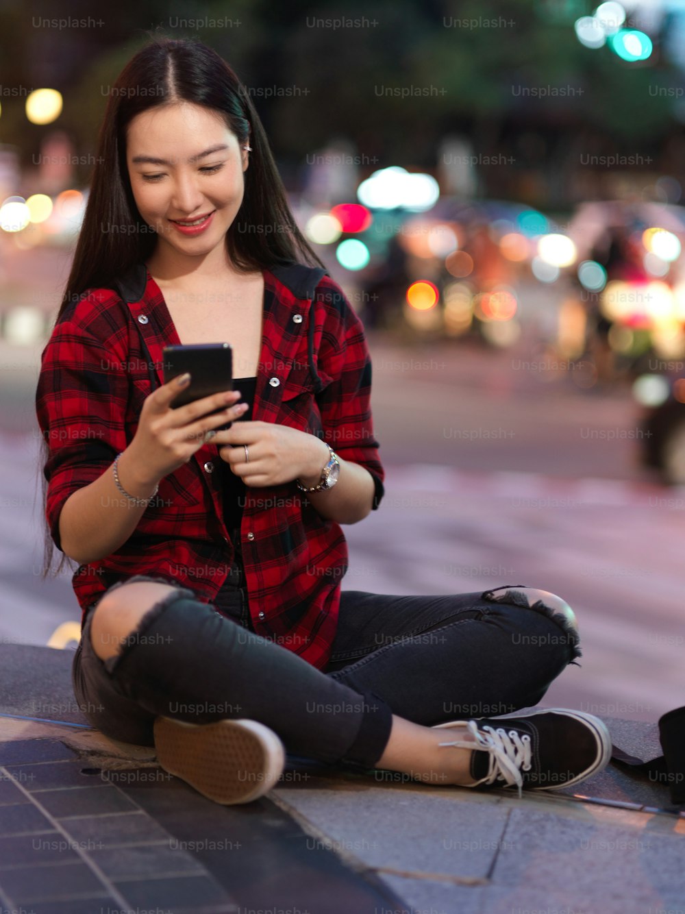 Portrait of happy hipster female sitting on stairs and using smartphone, chatting with friends, downloading application, updating social media, blurry city night lights in background