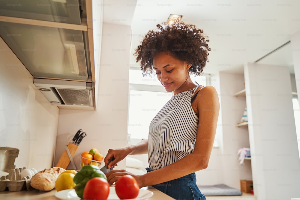 Smiling pleased curly-headed pretty lady dressed in casual clothes cutting vegetables with a chefs knife on the kitchen countertop