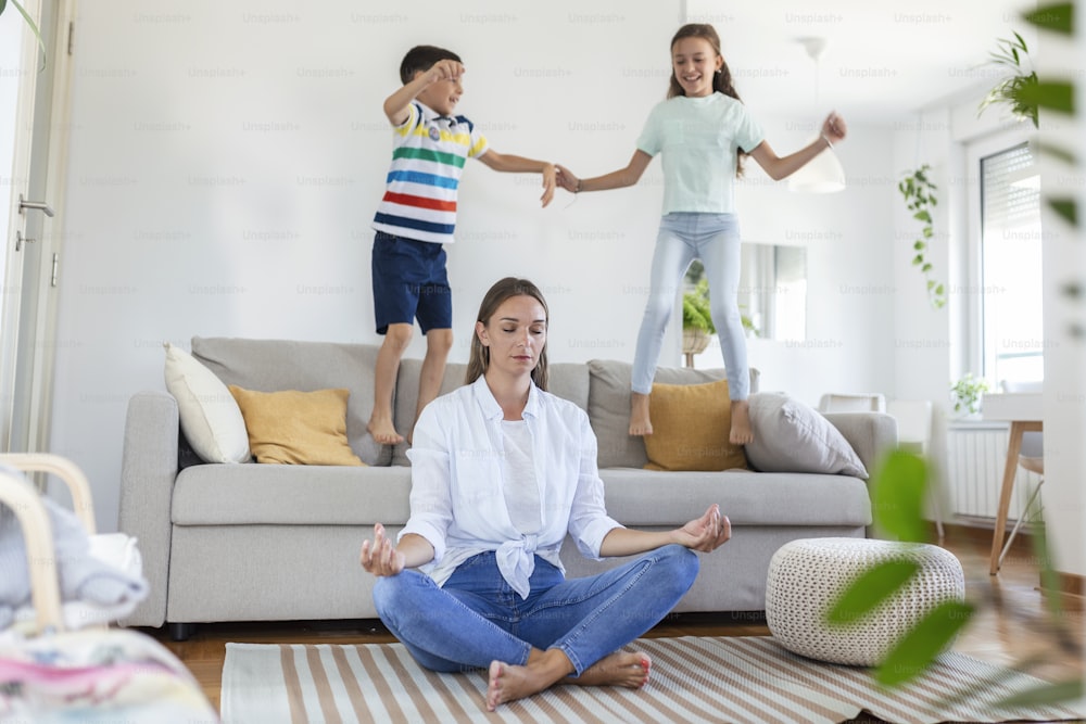 Young mother with closed eyes meditating in lotus pose on floor trying to save inner harmony while excited children jumping on sofa and screaming in light spacious living roo