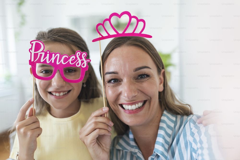 Funny family on a background of bright wall. Mother and her daughter girl with a paper accessories. Mom and child are holding paper crown on stick.