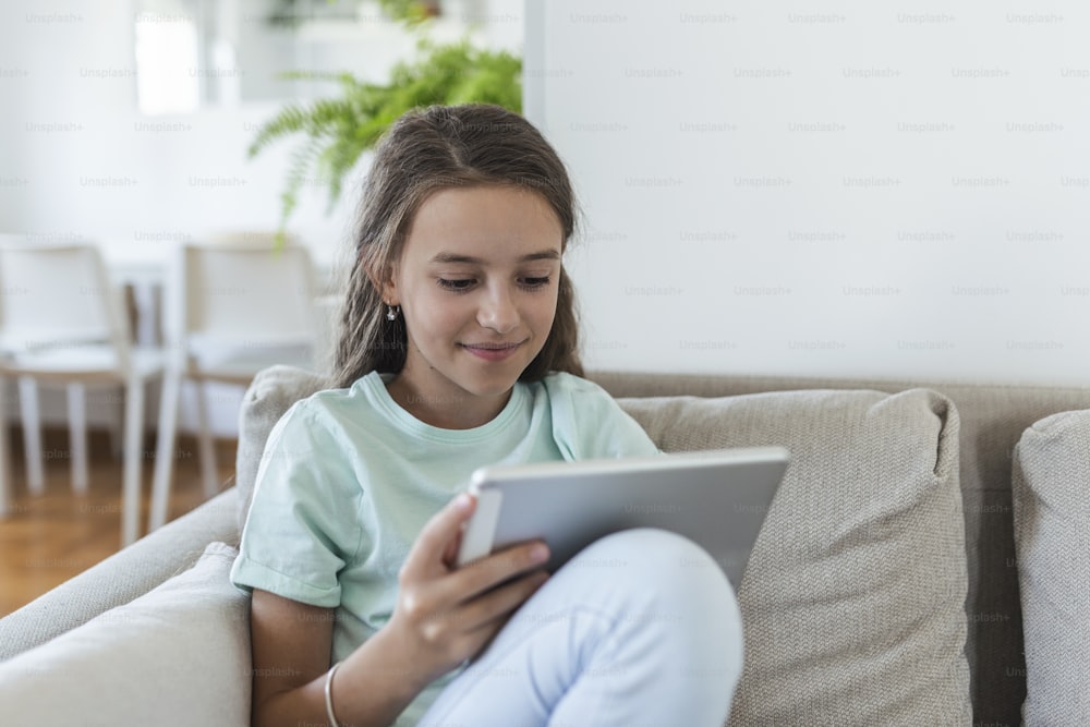 Mignon petit sourire de fille heureuse assis sur le canapé à l’aide d’un pad de tablette numérique à la recherche dans le salon à la maison. concept d’activité familiale.