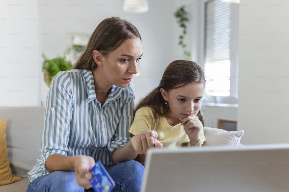 Cute small daughter rejoicing while smiling mother in casual clothes focusing on screen and using laptop for online shopping in light modern apartment