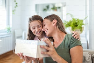 Young woman and girl at home celebrating mother's day sitting on sofa daughter hugging mother kissing cheek mom laughing joyful holding gift box