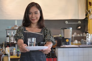 Eine schöne Barista-Frau hält eine Kaffeetasse und lächelt in die Kamera, während sie in der Nähe der Bartheke im Café steht.