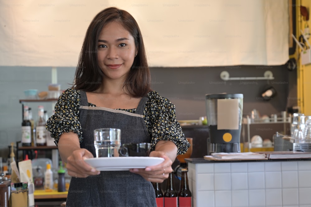 Beautiful female barista is holding coffee cup and smiling at camera while standing near the bar counter in cafe.