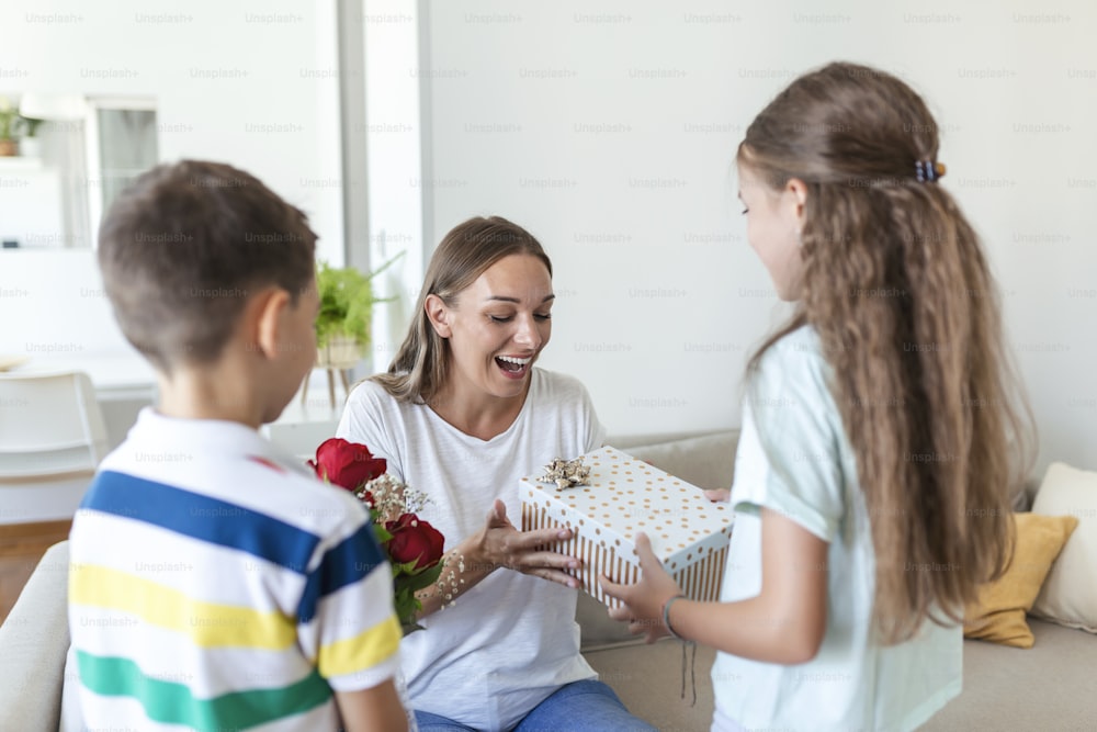 Happy children giving gift an flowers to mother. Happy Mothers Day! Children boy and girl congratulate smiling mother, give her flowers bouquet of roses and a gift box during holiday celebration