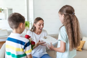 Happy children giving gift an flowers to mother. Happy Mothers Day! Children boy and girl congratulate smiling mother, give her flowers bouquet of roses and a gift box during holiday celebration